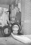 Two men standing outside of the ladie's restroom of a Gulf Service Station next to two tires. by William Garber