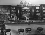Inside of Everly Drug Store, view of bar stools at a soda counter top facing a mirror and giant Coca-Cola ad. Shelves of magazines can be seen to the left and shelves of medicines or hygienic products in the mirror. by William Garber