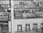 Inside of a store, view of a woman standing behind the counter. by William Garber