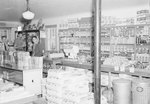 Inside of a store, view of shelving stocked with various food items, and four men standing to the far left. by William Garber