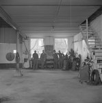 Distant view of four men standing in the back of a large shop, garage, or warehouse room by William Garber