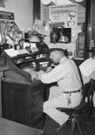 Side view of a man in a work uniform working at a cluttered desk by William Garber