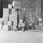 A young boy sitting on the floor in front of a tall stack of boxes by William Garber