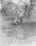 Two men outside either laying down pipes or tubing, or taking measurements by William Garber