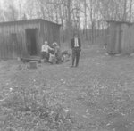 Three men standing and sitting outside of two wooden sheds or shacks by William Garber