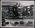 Inside of a business, probably a general or drug store. View of a counter with bar stools and a large Coca-Cola advertisement by William Garber