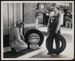 Two men, one standing and one crouching, outside of an automotive shop next to an advertisement for Gulf tires by William Garber