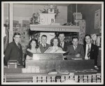John Deere Day, five men and two women posing behind a counter that is advertising John Deere repair parts by William Garber