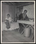 Two African American men sitting inside of small wooden home or cabin; the one man is posing at a table while writing, while the other poses from the bed, where he is reading by William Garber