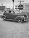 A private automobile parked next to a Coca-Cola advertisement and sign that says "Happy Motoring" by William Garber
