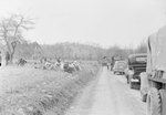 A group of CCC workers sitting in an overgrown field facing a dirt road that is lined with various automobiles by William Garber