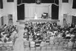 New Market High School, view from the back of the auditorium, where a woman and a man are playing the violin and piano on stage by William Garber