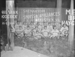 Team photo of the Quicksburg men's baseball team, with the fence of a baseball field advertising Woodstock businesses in the background by William Garber