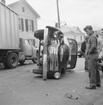Front-end view of an automobile that is laying on its side on a residential street. A tractor-trailer is in the background. by William Garber