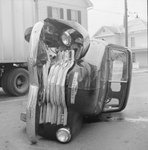 Front-end view of a vehicle that is laying on its side on a residential street. A tractor-trailer is in the background. by William Garber